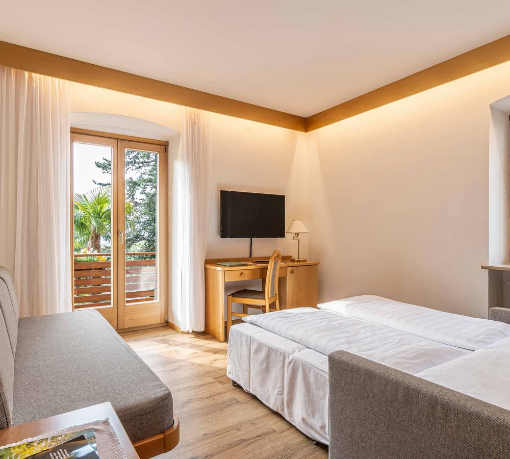 Helles Hotelzimmer mit zwei Einzelbetten, Sofa, Schreibtisch, TV und Balkon mit Blick ins Grüne