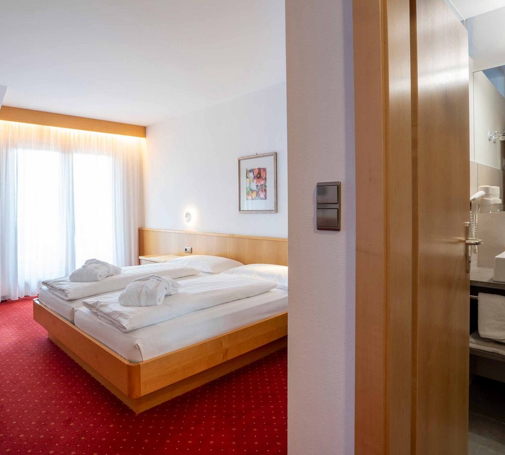 Helles Hotelzimmer mit rotem Teppich, Doppelbett, Tisch mit Stühlen, Fenster mit weißen Vorhängen und angrenzendem Badezimmer