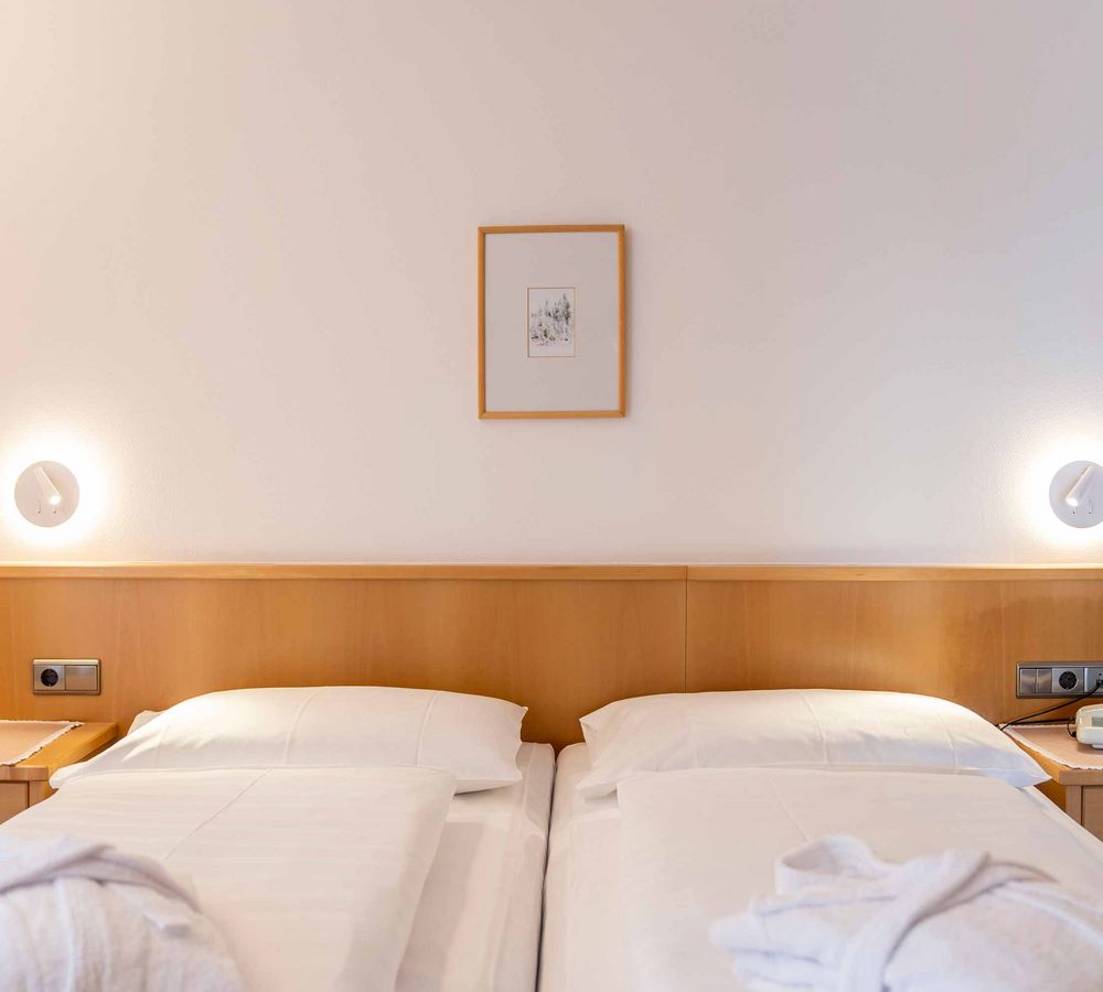 Zwei nebeneinander stehende Einzelbetten mit weißen Bettlaken und Kopfkissen, je ein Nachttisch mit Lampe an der Wand und ein kleines Bild über den Betten.