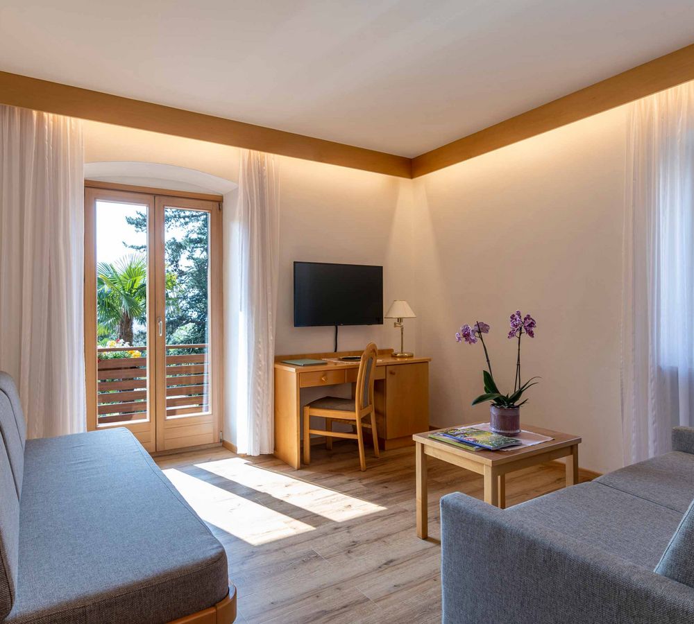 Helles Wohnzimmer mit zwei grauen Sofas, Holztisch mit Blume, Schreibtisch mit Stuhl und Fernseher, große Fenster mit Vorhängen und Balkonblick