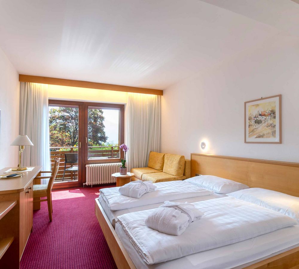 Helles Hotelzimmer mit Doppelbett, rotem Teppich, Sofa, Schreibtisch, Fernseher und Balkon mit Blick auf Bäume.