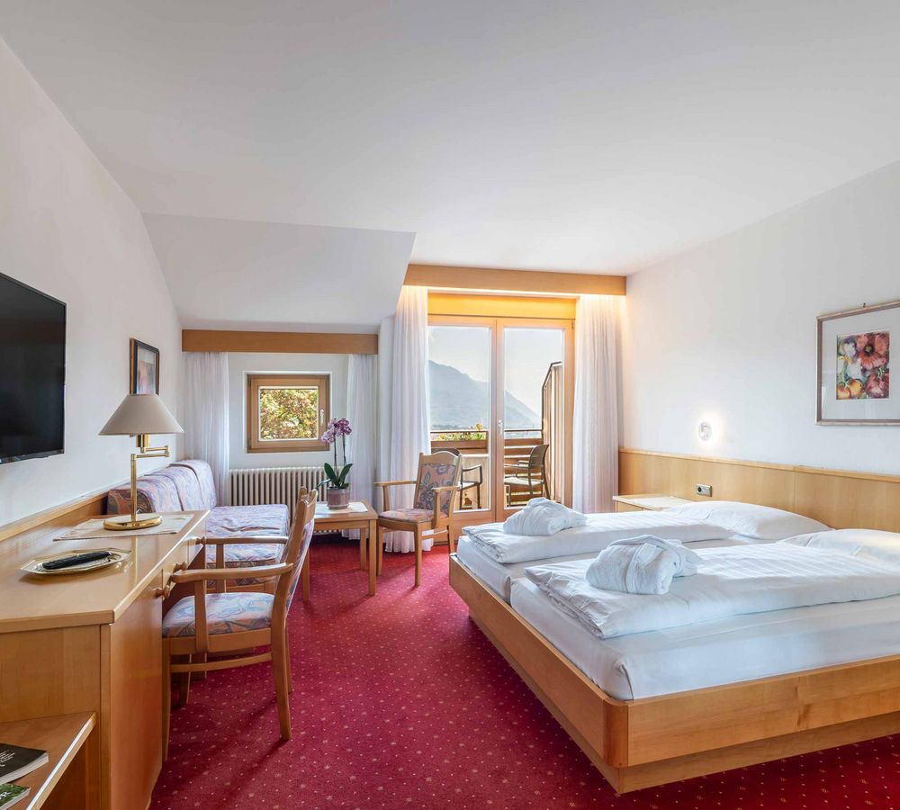 Helles Hotelzimmer mit Doppelbett, roten Teppichboden, Holzmöbeln und Balkon mit Bergblick