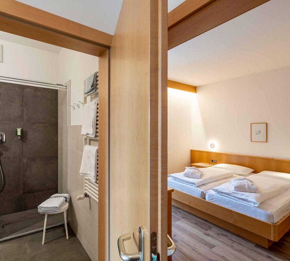 Blick durch eine geöffnete Holztür auf ein modernes Badezimmer mit Dusche und ein helles Schlafzimmer mit zwei Einzelbetten.