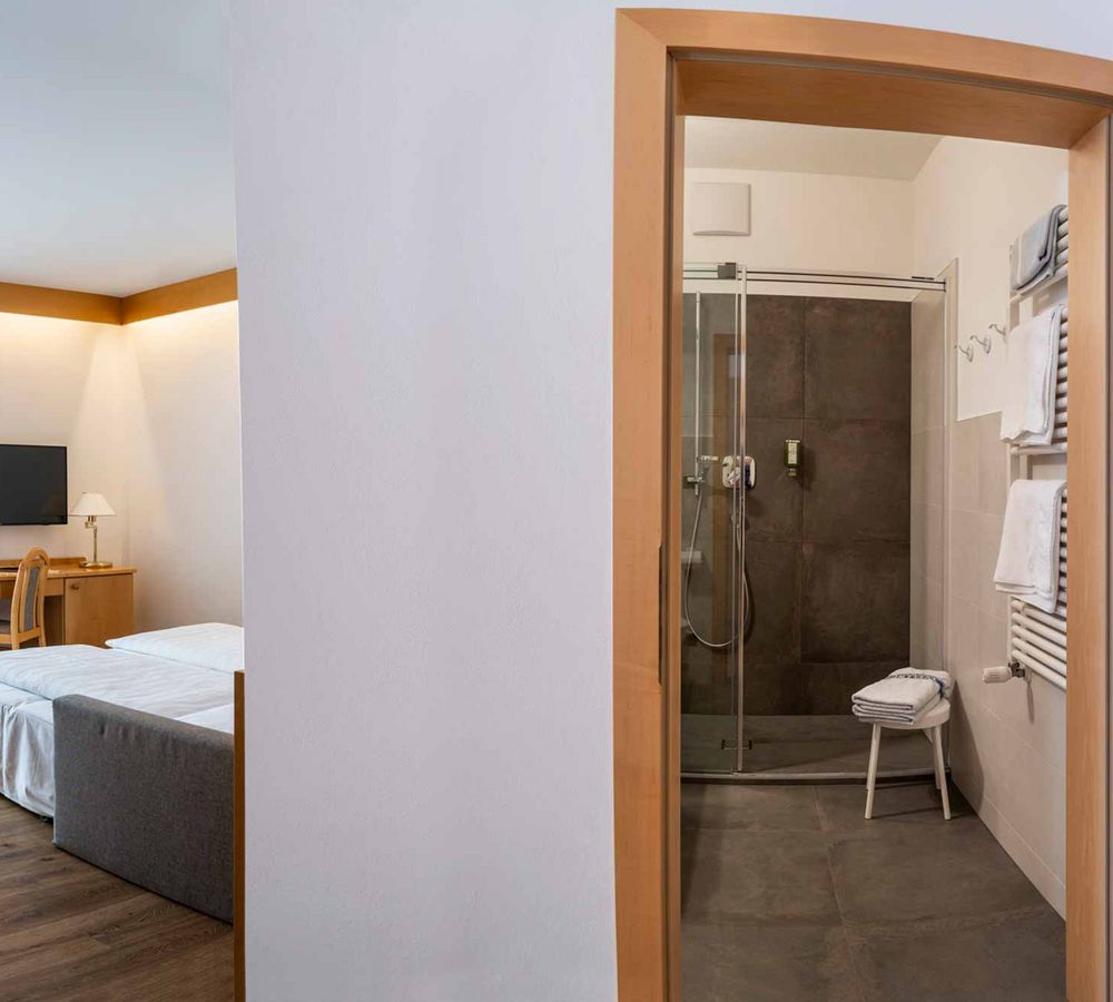 Blick in ein helles Hotelzimmer mit zwei Betten, Sitzbereich, Schreibtisch, Fernseher und angrenzendem Badezimmer mit Dusche