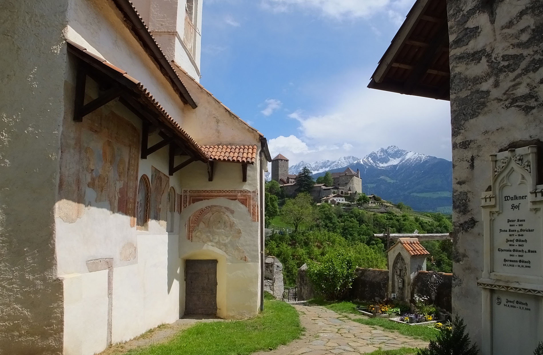 Wanderweg zwischen historischen Gebäuden mit bemalten Wänden, im Hintergrund ein Schloss auf einem Hügel und schneebedeckte Berge unter blauem Himmel.