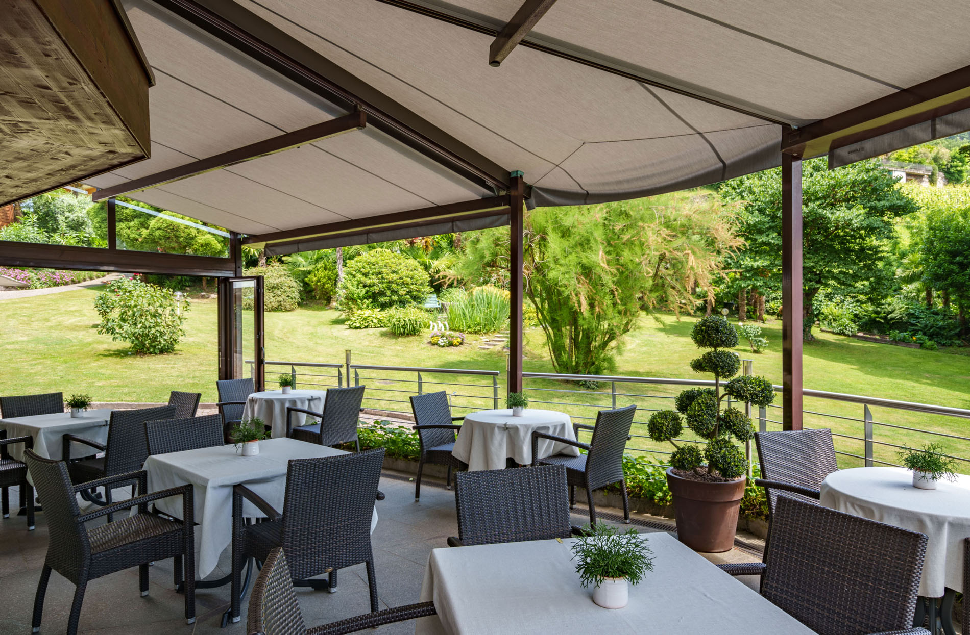 Überdachte Terrasse mit mehreren gedeckten Tischen und Stühlen vor einem Garten mit grünen Pflanzen und Büschen Überdachte Terrasse mit mehreren gedeckten Tischen und Stühlen vor einem Garten mit grünen Pflanzen und Büschen