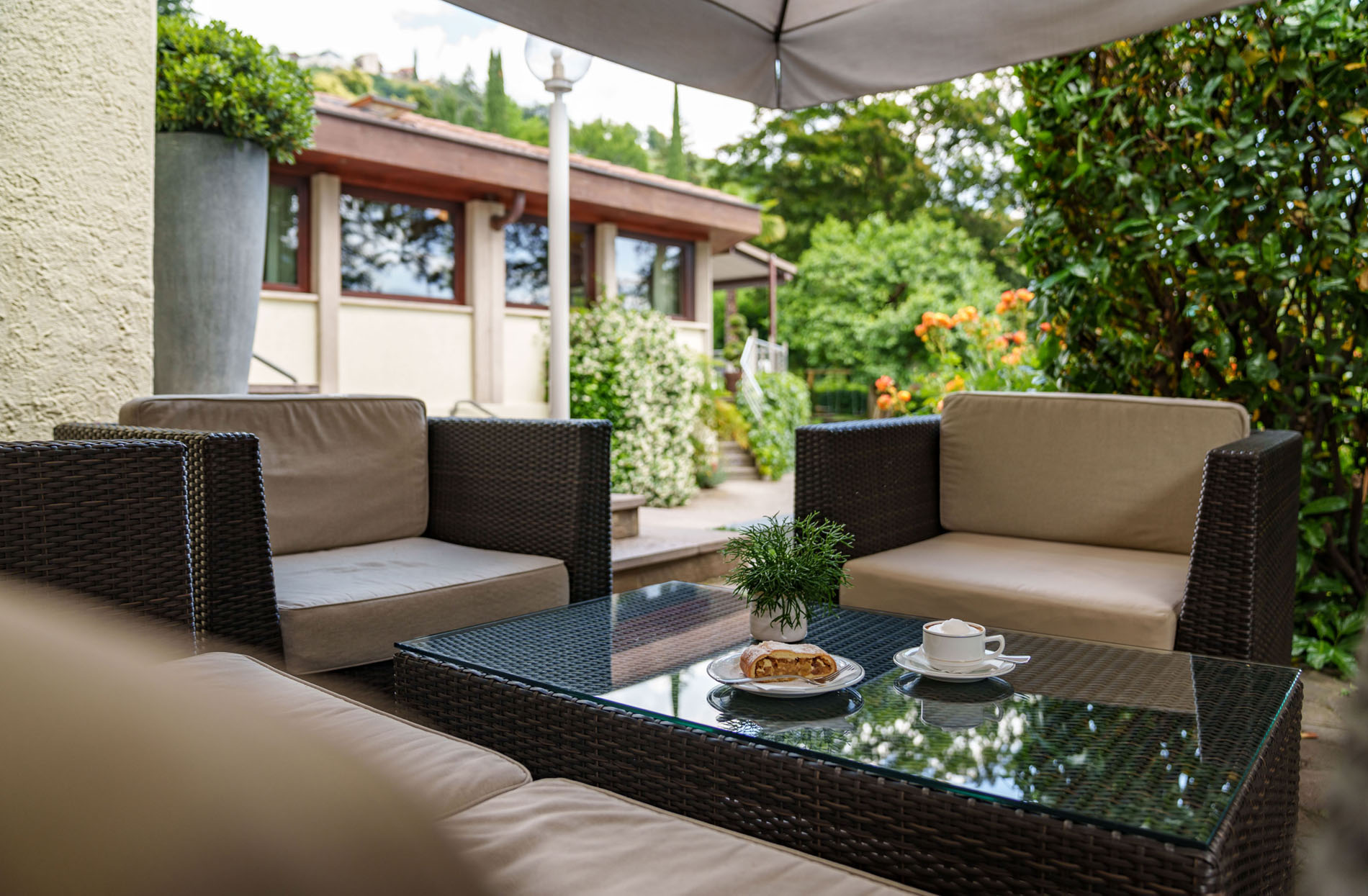 Gemütliche Terrasse mit Rattanmöbeln, Kaffeetasse, Stück Kuchen und grünem Pflanzenarrangement auf dem Glastisch Gemütliche Terrasse mit Rattanmöbeln, Kaffeetasse, Stück Kuchen und grünem Pflanzenarrangement auf dem Glastisch