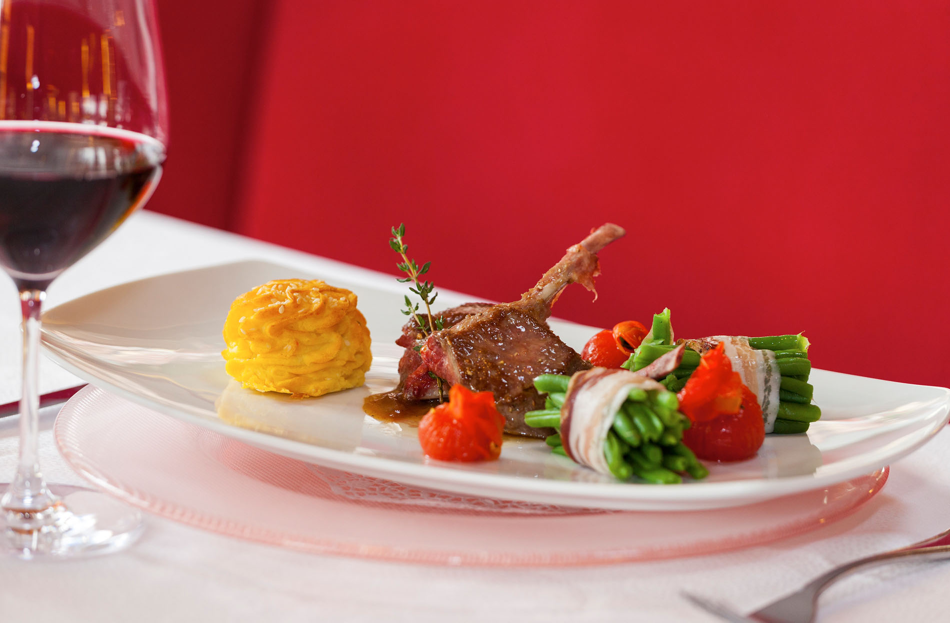 Elegantes Gericht mit Lammkotelett, grünen Bohnen in Speck, Tomaten und einer Sauce, serviert auf weißem Teller mit Glas Rotwein vor rotem Hintergrund. Elegantes Gericht mit Lammkotelett, grünen Bohnen in Speck, Tomaten und einer Sauce, serviert auf weißem Teller mit Glas Rotwein vor rotem Hintergrund.