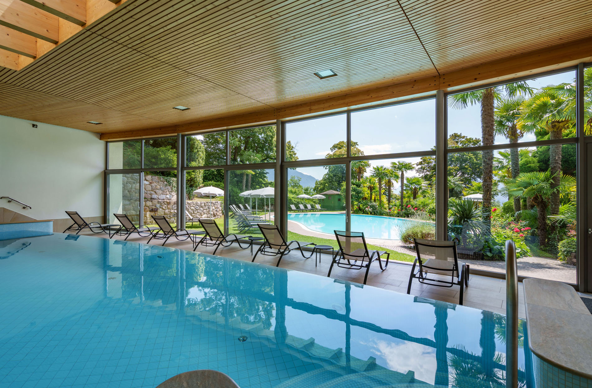 Innenpool mit Liegestühlen vor großen Glasfenstern, durch die ein Außenpool und Palmen zu sehen sind