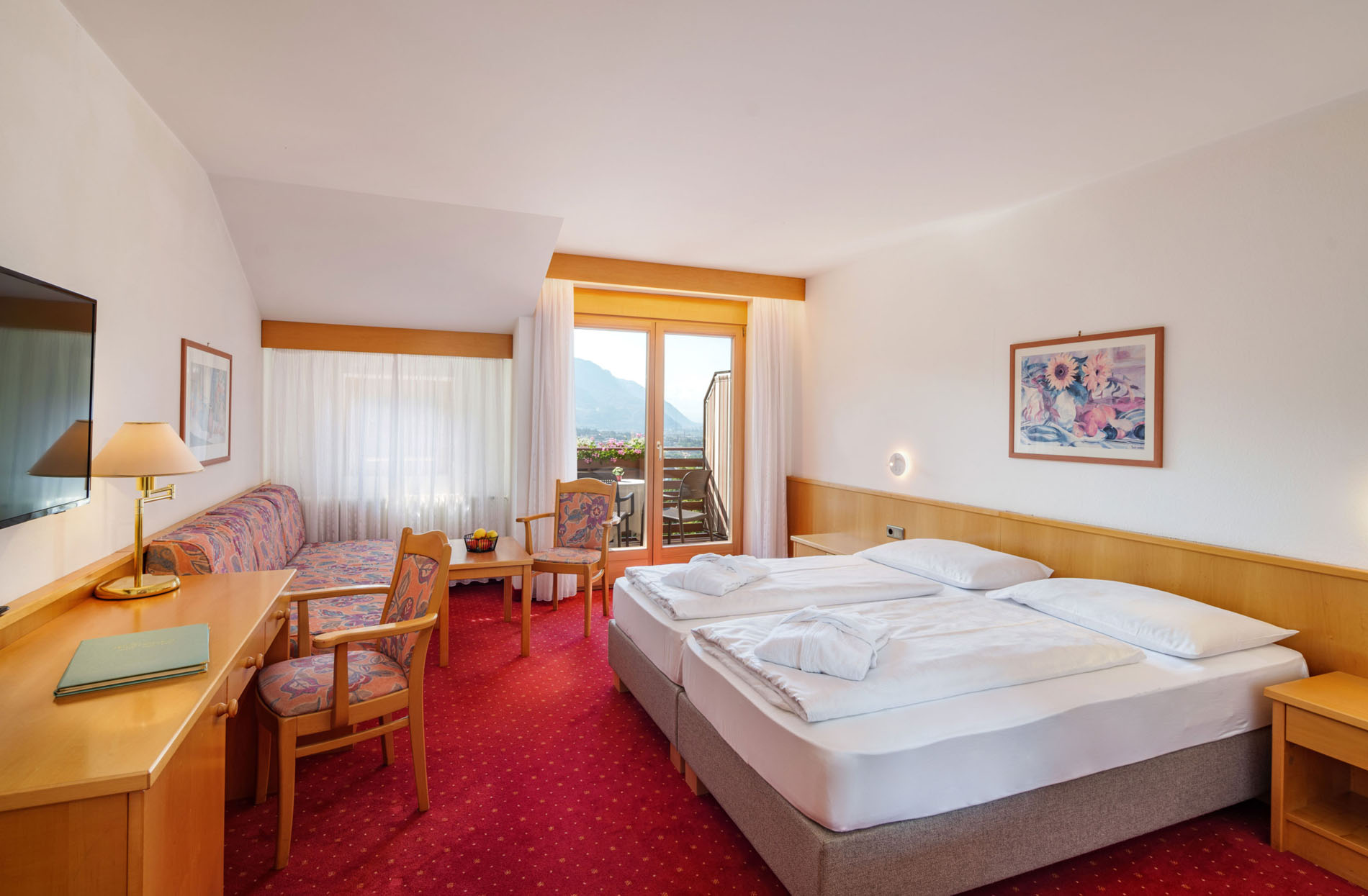 Helles Hotelzimmer mit zwei Einzelbetten, rotem Teppich, Holzmöbeln und Balkon mit Blick auf Berge.