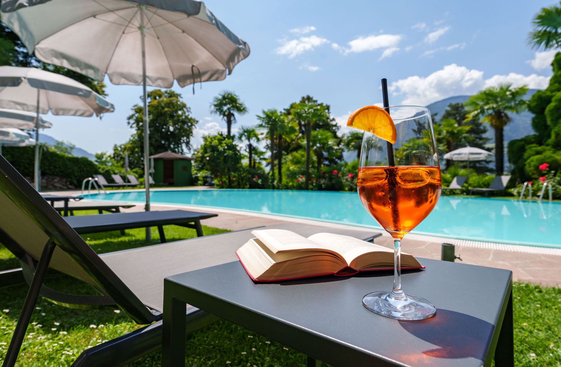 Cocktailglas mit Orangenscheibe neben einem aufgeschlagenen Buch auf einem Tisch am Pool mit Sonnenliegen und Sonnenschirmen