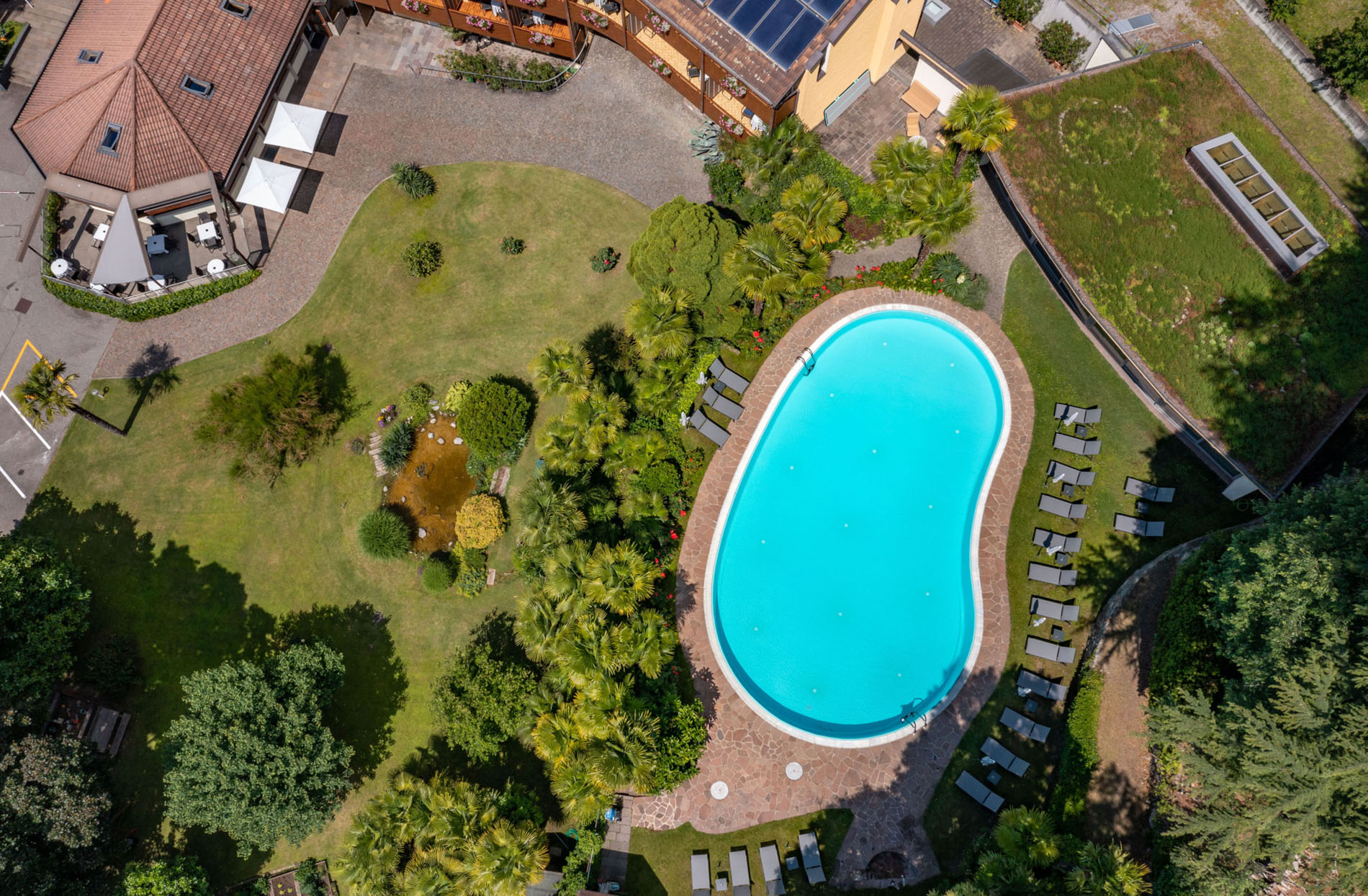Luftaufnahme eines Gartens mit Swimmingpool, umgeben von Liegestühlen, Palmen und einem Teich, neben Gebäuden mit begrünten Dächern. Luftaufnahme eines Gartens mit Swimmingpool, umgeben von Liegestühlen, Palmen und einem Teich, neben Gebäuden mit begrünten Dächern.