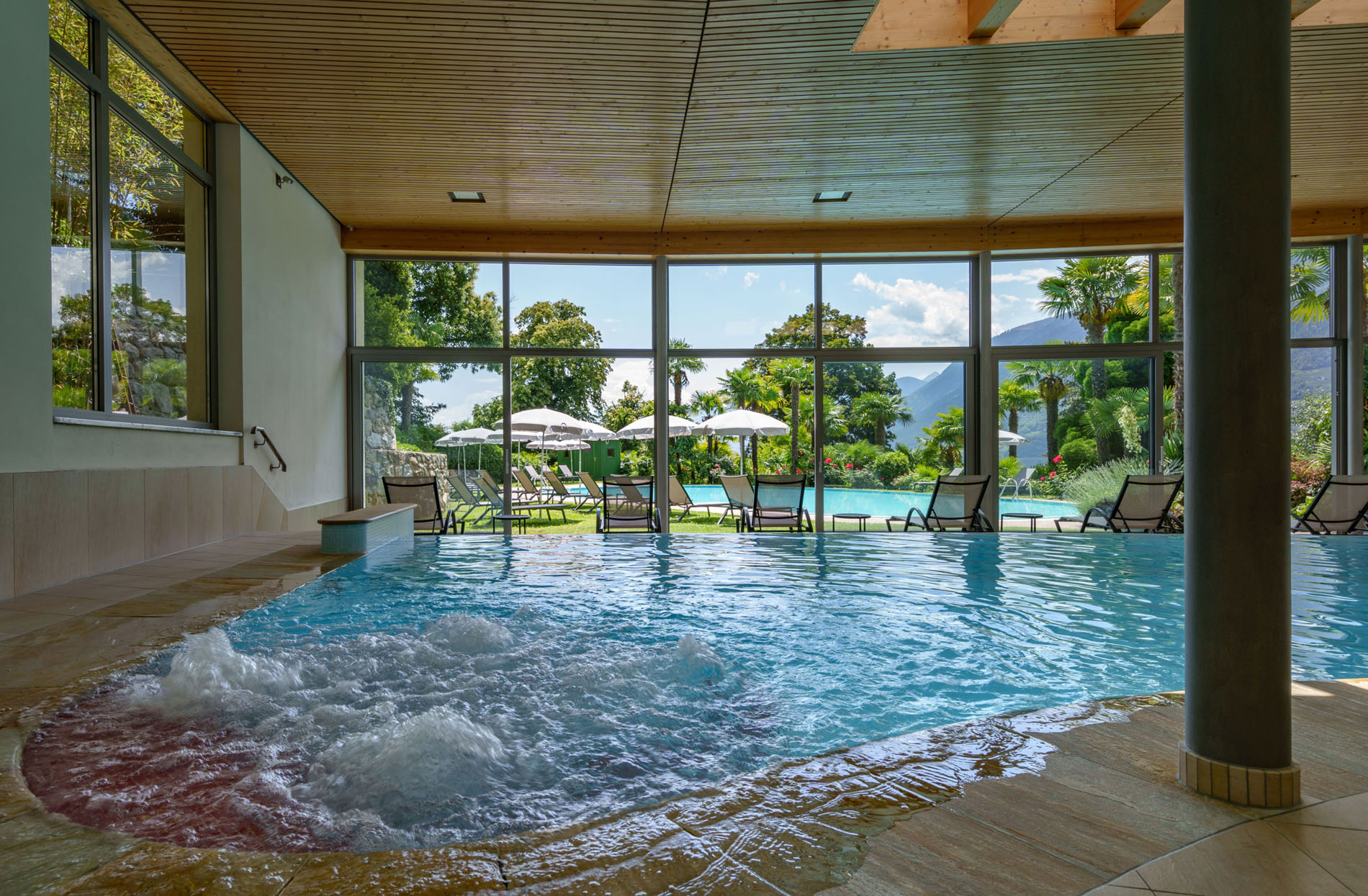 Innenblick auf den Whirlpool und Poolbereich mit großen Fenstern, durch die Liegestühle, Sonnenschirme und Garten mit Bäumen und Bergen im Hintergrund sichtbar sind. Innenblick auf den Whirlpool und Poolbereich mit großen Fenstern, durch die Liegestühle, Sonnenschirme und Garten mit Bäumen und Bergen im Hintergrund sichtbar sind.